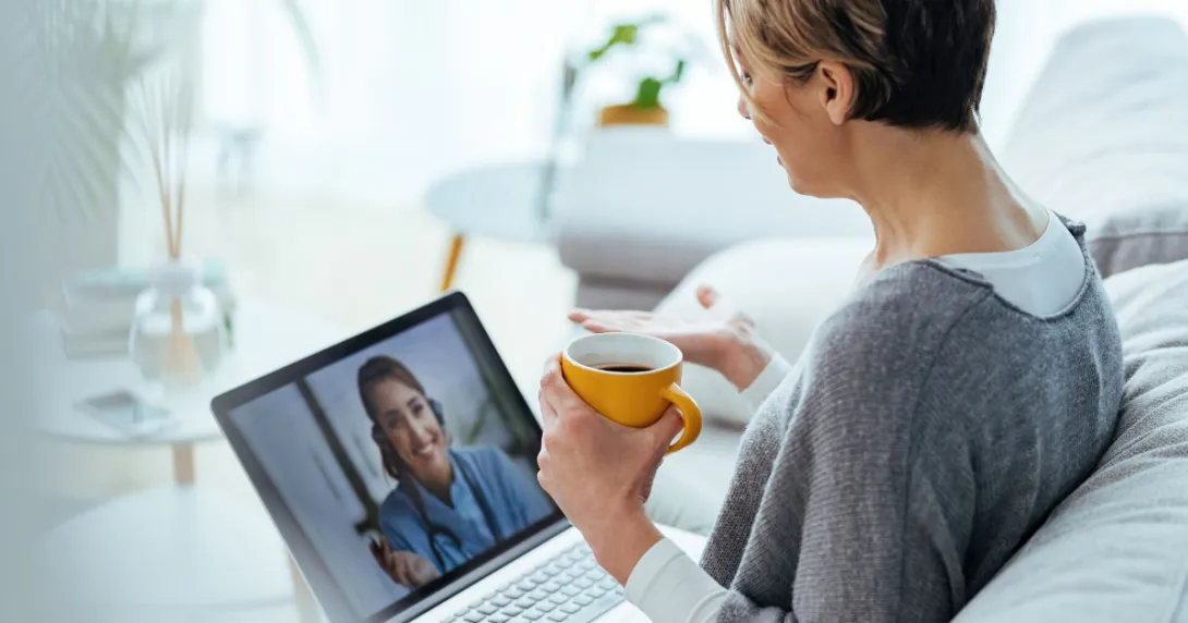 Person sitting on a couch holding a coffee cup while talking to a healthcare professional via a laptop Person sitting on a couch holding a coffee cup while talking to a healthcare professional via a laptop