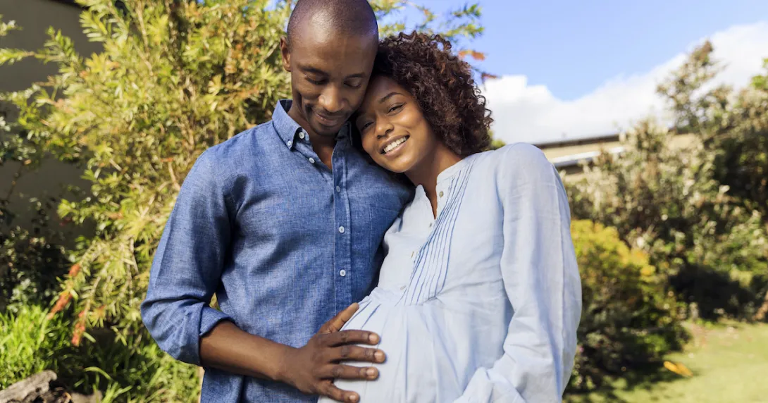 
  A pregnant person standing in a garden next to another person with their hand on the pregnant person's stomach
