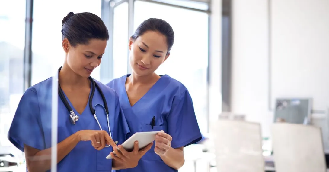 Two healthcare providers wearing blue scrubs and looking at a tablet Two healthcare providers wearing blue scrubs and looking at a tablet