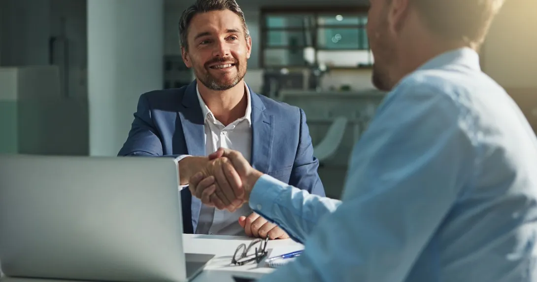 Two people sitting on opposite sides of a desk and shaking hands