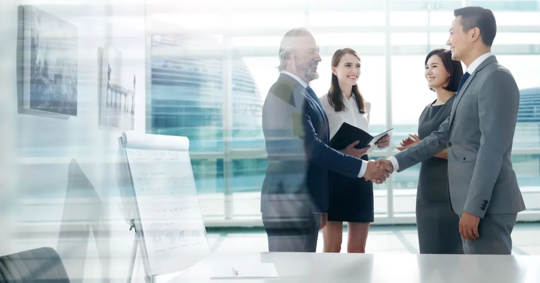 Four people standing in front of a large window with two of them shaking hands and the other two watching