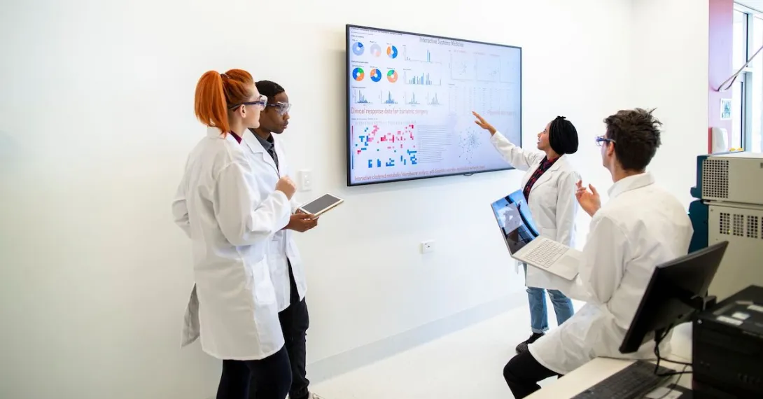 Professional team of medical scientists in a laboratory gathered around a wall monitor discussing charts and data