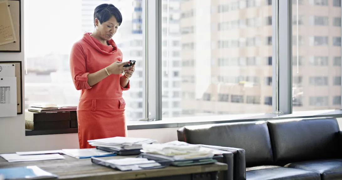 Businesswoman standing in office looking at smart phone smiling